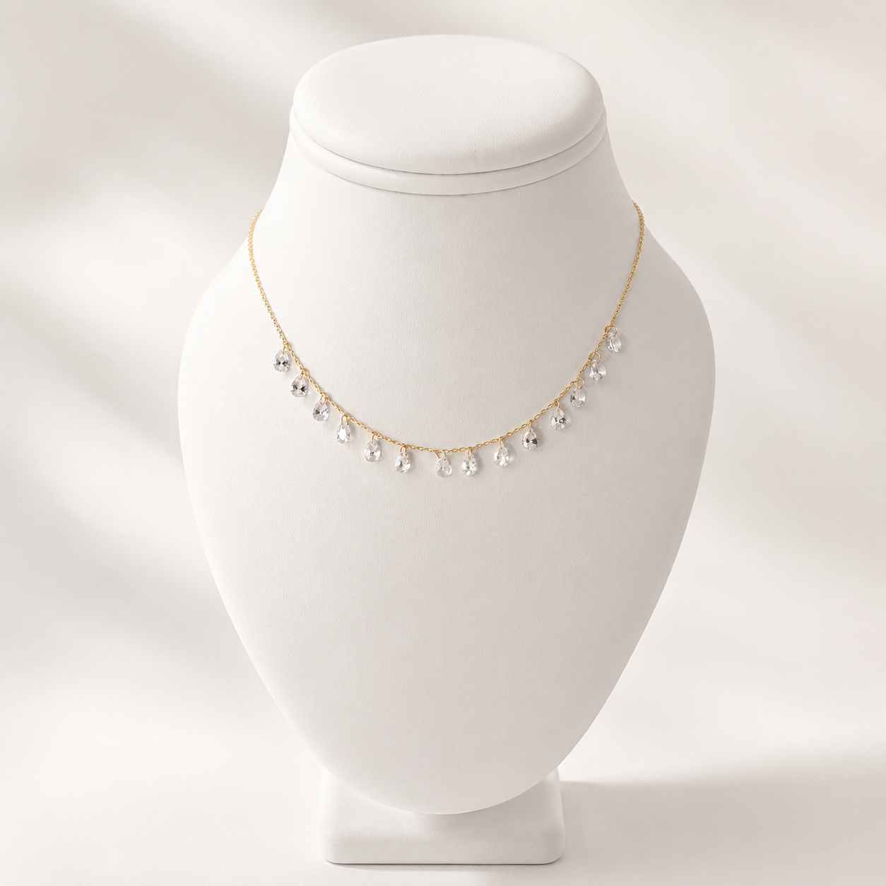 A delicate gold chain necklace with small clear teardrop-shaped crystals, displayed on a white jewelry bust against a soft white background.