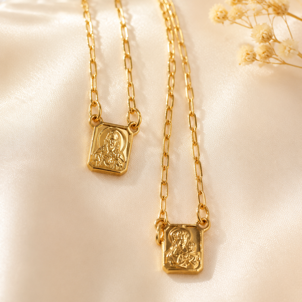 Two gold necklaces with rectangular pendants featuring embossed religious figures, displayed on a soft cream satin fabric with dried flowers in the corner.