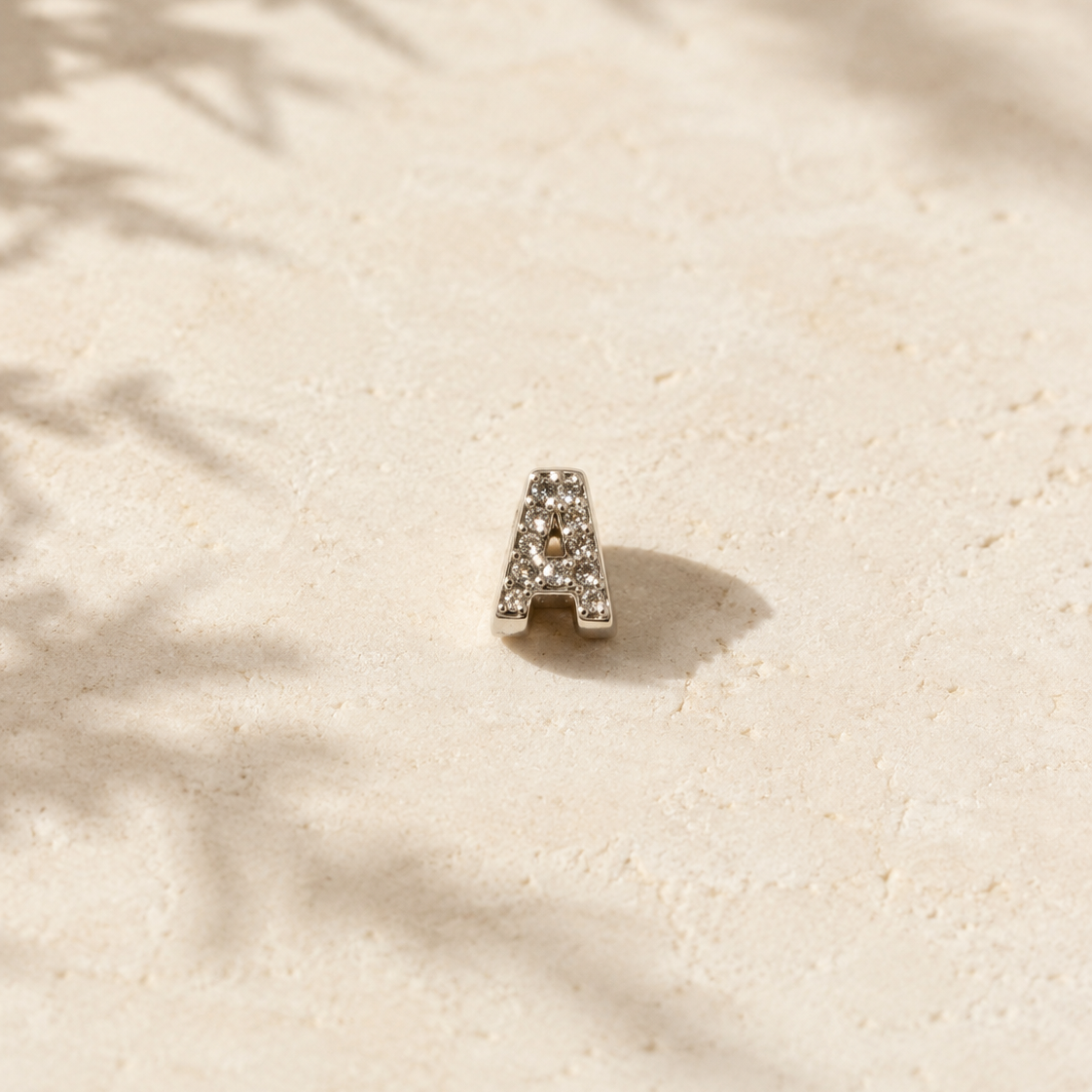 Silver letter 'A' charm on a textured beige surface with leaf shadows