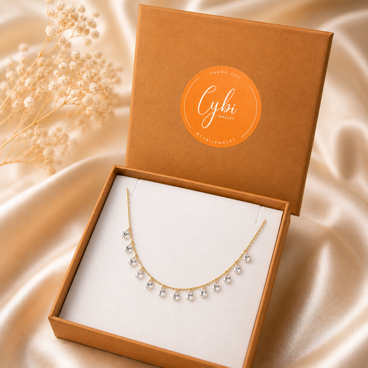 A delicate gold necklace with clear crystal-like beads draped over an open brown jewelry box lined with white fabric. The box lid features a circular orange sticker reading 'Cybi Jewellery Thank You 
