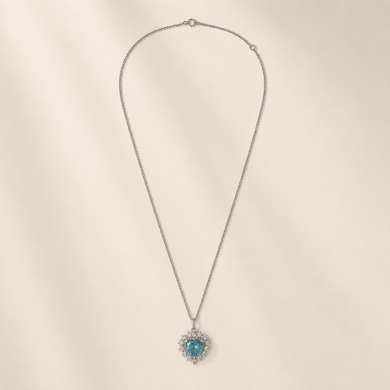A silver-toned chain necklace with a heart-shaped pendant featuring a central turquoise-blue stone surrounded by small clear stones, displayed on a soft beige background.