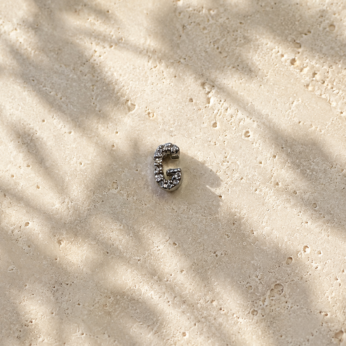 Silver letter 'G' charm on sandy surface with leaf shadows