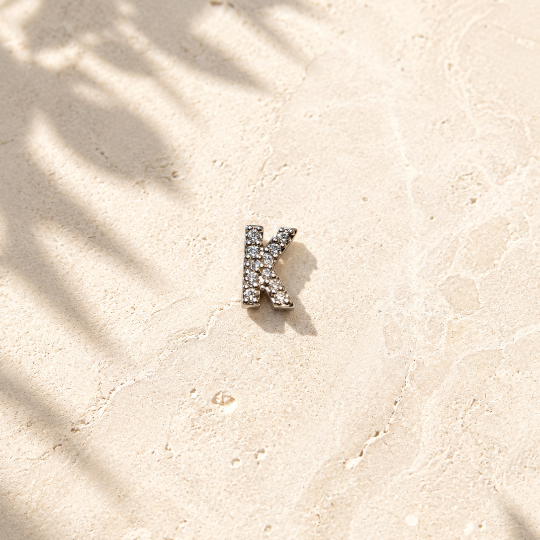 Silver letter 'K' earring on a sandy surface with palm leaf shadows