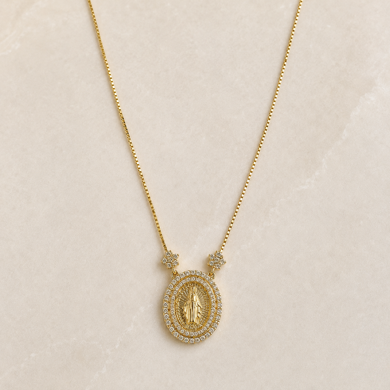 A gold necklace with a circular pendant featuring a religious figure surrounded by small clear stones, displayed on a light beige textured background.