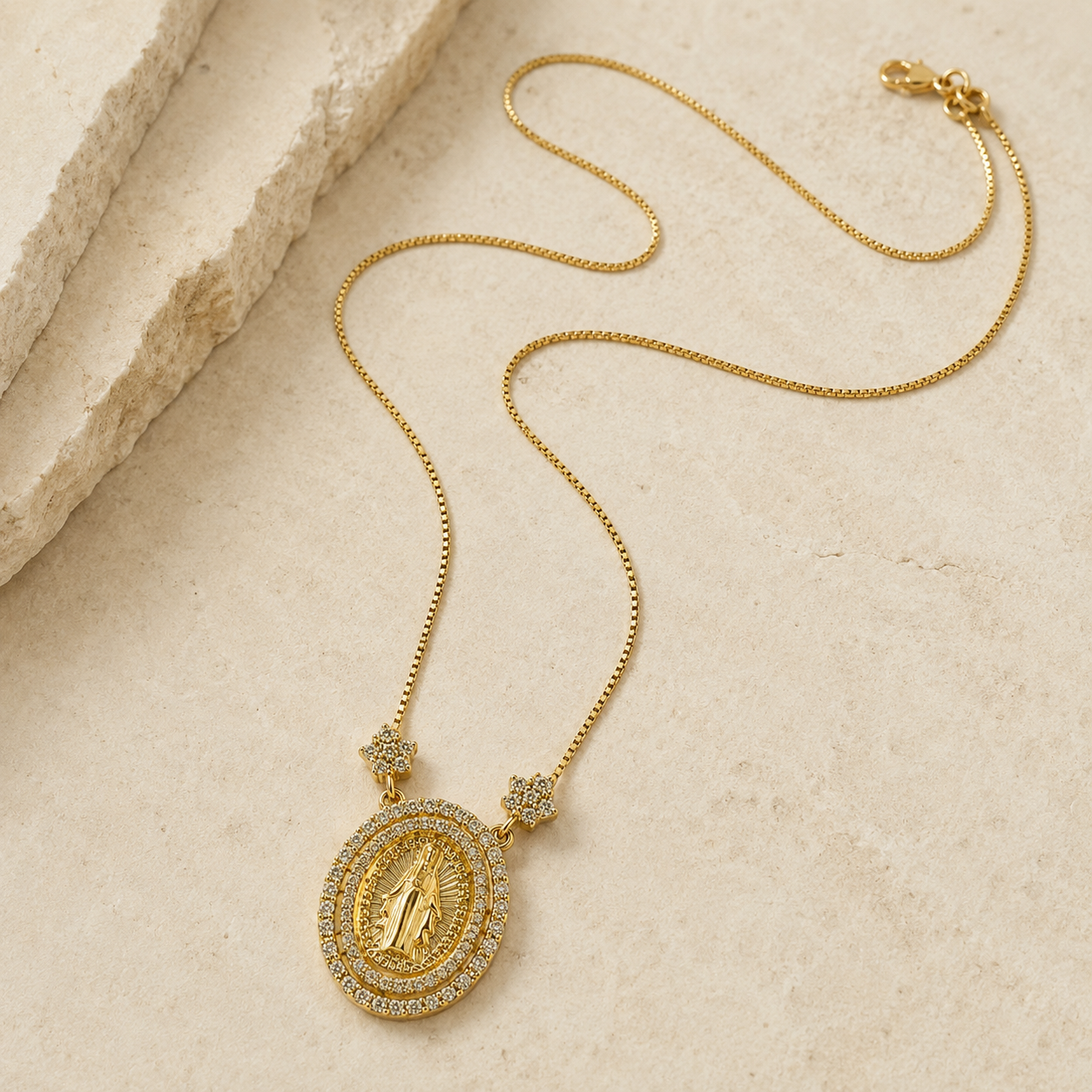 A gold chain necklace with an oval religious pendant featuring a gold figure surrounded by small clear stones, displayed on a light beige textured stone surface.