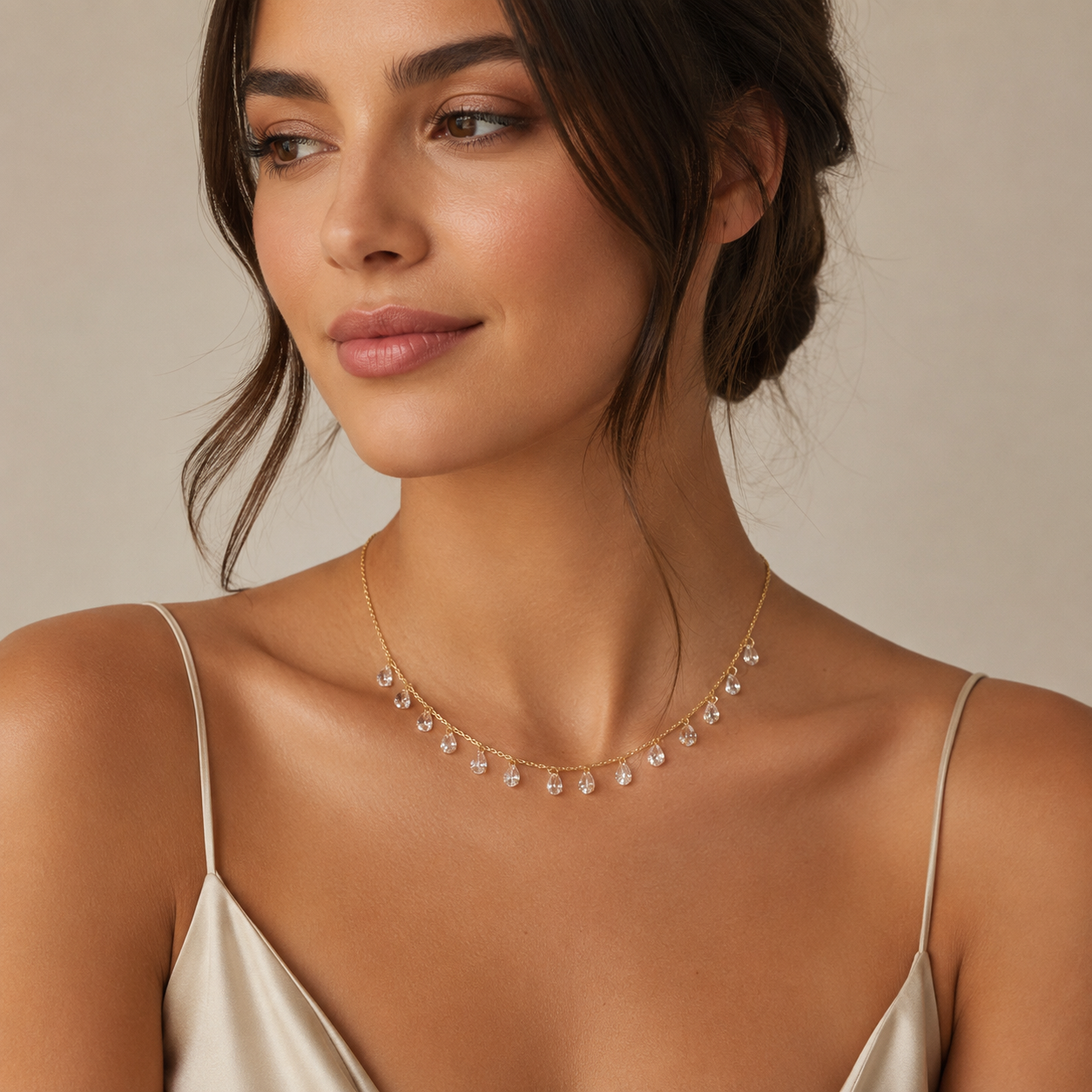 A woman wearing a delicate gold necklace with small clear crystal drops, photographed against a soft beige studio background.